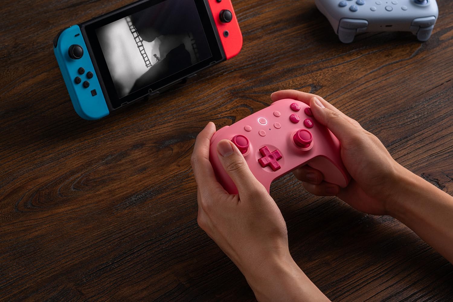 Hands holding pink gaming controller with consoles.